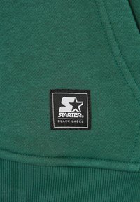 Zielona bluza z textured materiału, z czarną kwadratową metką, na której znajduje się widoczny napis "STARTER" oraz logo gwiazdki.