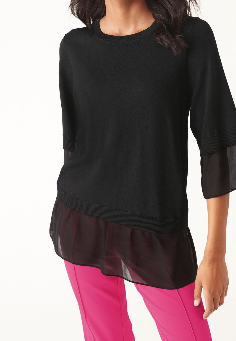 Donna che indossa un top nero a strati con orlo e maniche trasparenti, abbinato a pantaloni rosa brillante, mostrata dalla gola ai fianchi.