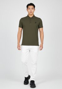 Junger Mann, der ein olivgrünes Poloshirt, weiße Cargo-Hosen und schwarze Sneaker trägt, steht und blickt frontal vor einem einfarbigen Hintergrund.