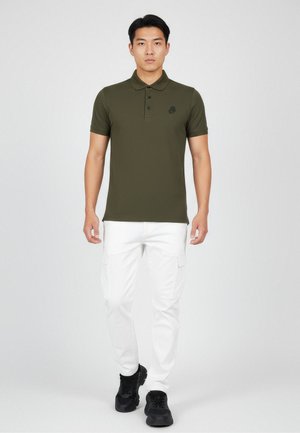 Junger Mann, der ein olivgrünes Poloshirt, weiße Cargo-Hosen und schwarze Sneaker trägt, steht und blickt frontal vor einem einfarbigen Hintergrund.