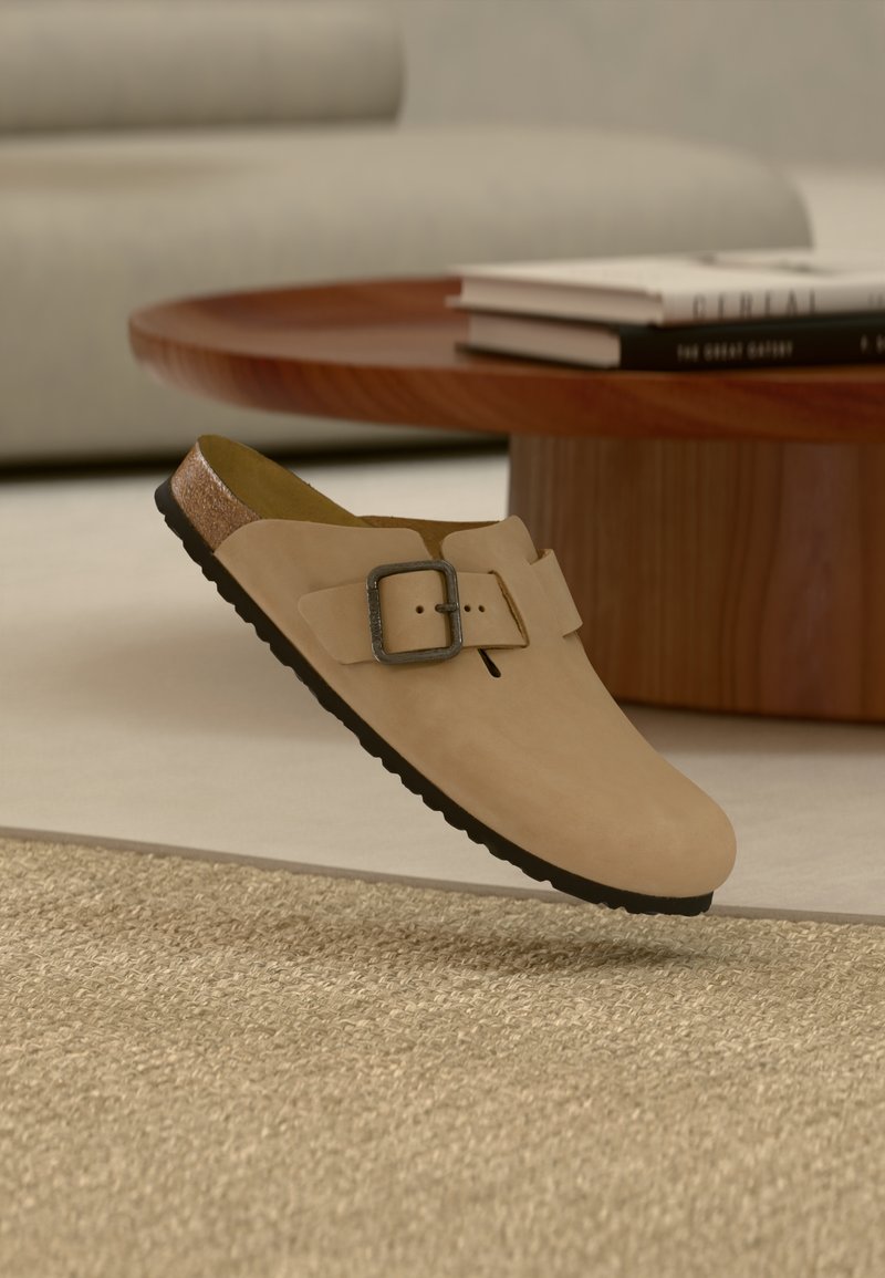 Μπεζ παπούτσι τύπου clog με λουράκι με ρυθμιζόμενο πόμολο πάνω σε υφαντό χαλί, με στρογγυλό ξύλινο τραπεζάκι καφέ και βιβλία στο φόντο.