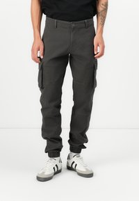 Pantalons cargo en coton gris foncé avec poches latérales, jambes fuselées et poignets élastiques. Portés avec des baskets blanches et grises avec des rayures noires.