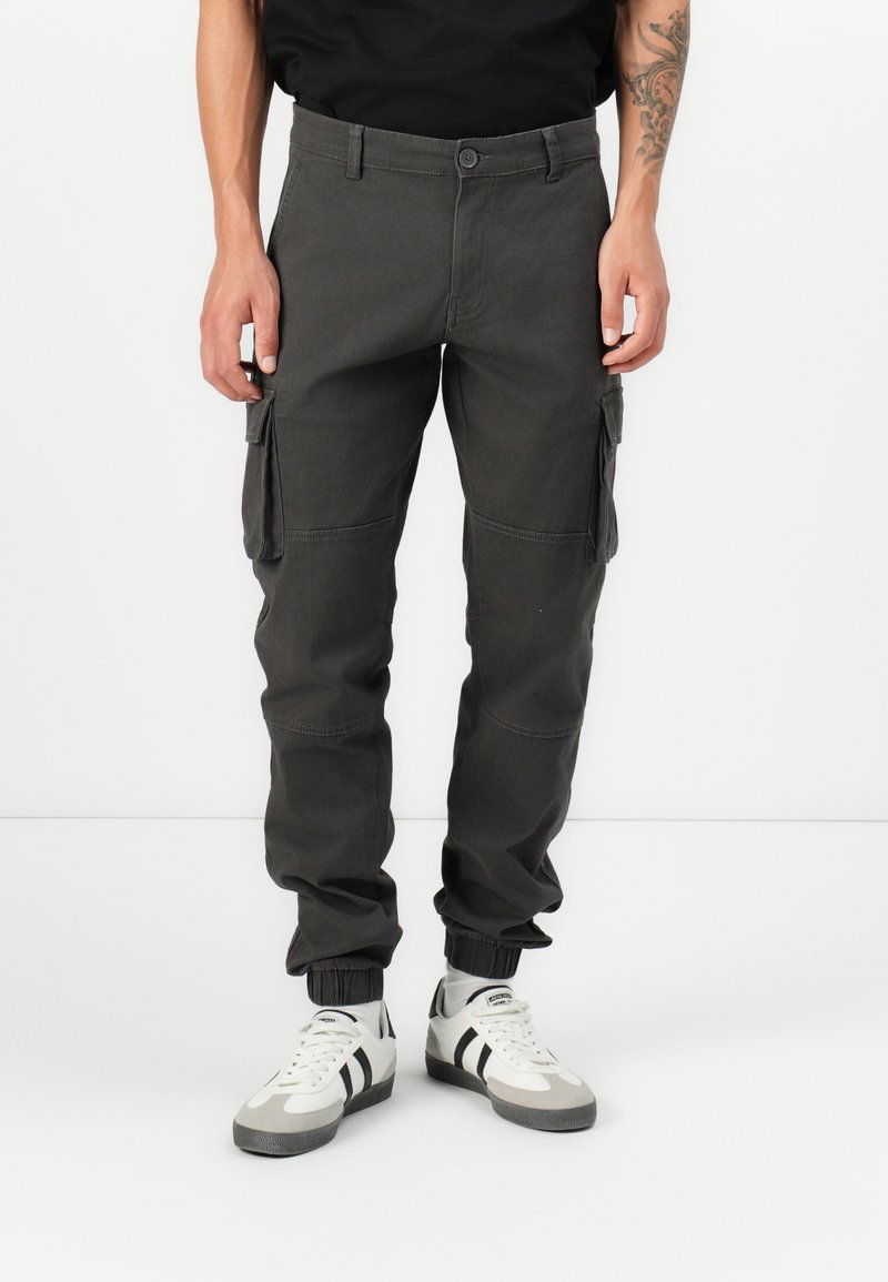 Pantalons cargo en coton gris foncé avec poches latérales, jambes fuselées et poignets élastiques. Portés avec des baskets blanches et grises avec des rayures noires.