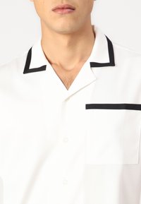 White short-sleeve shirt with a black piped collar and pocket. Smooth fabric, button placket, and a relaxed fit.