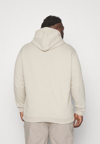 Felpa beige con cappuccio regolabile, vestibilità rilassata, polsini e vita a costine, e tessuto morbido, mostrata da dietro.