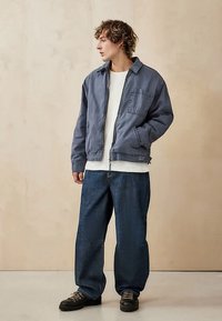 Młody mężczyzna w niebieskiej bluzie na zamek, białej koszuli, luźnych ciemnych dżinsach i czarnych butach, stojący z jedną ręką w kieszeni, oparty o beżową ścianę.