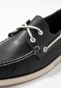 Zapato de barco de cuero negro con costuras en blanco que contrastan, cordones grises y suela de goma. Presenta ojales redondeados y una textura suave.
