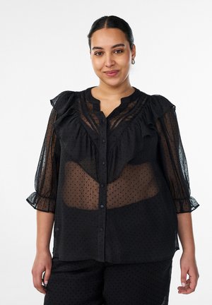 Femme aux cheveux foncés en chignon bas portant un chemisier noir transparent avec des volants et des pois, associé à un pantalon noir assorti, sur fond blanc.
