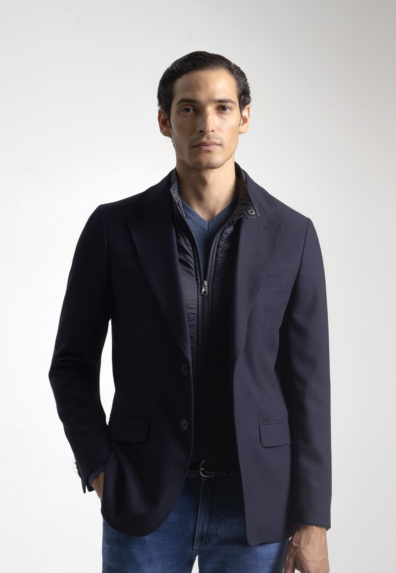 Blazer naval con un acabado texturizado superpuesto a una chaqueta con cremallera azul marino. Cuenta con un cuello con muescas, botones y un ajuste moderno combinado con unos vaqueros.