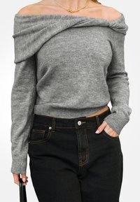 Szary, sweter typu off-the-shoulder o teksturowanym splocie, połączony z wysokimi czarnymi dżinsami z złotymi przeszyciami i zapięciem na guziki z przodu.