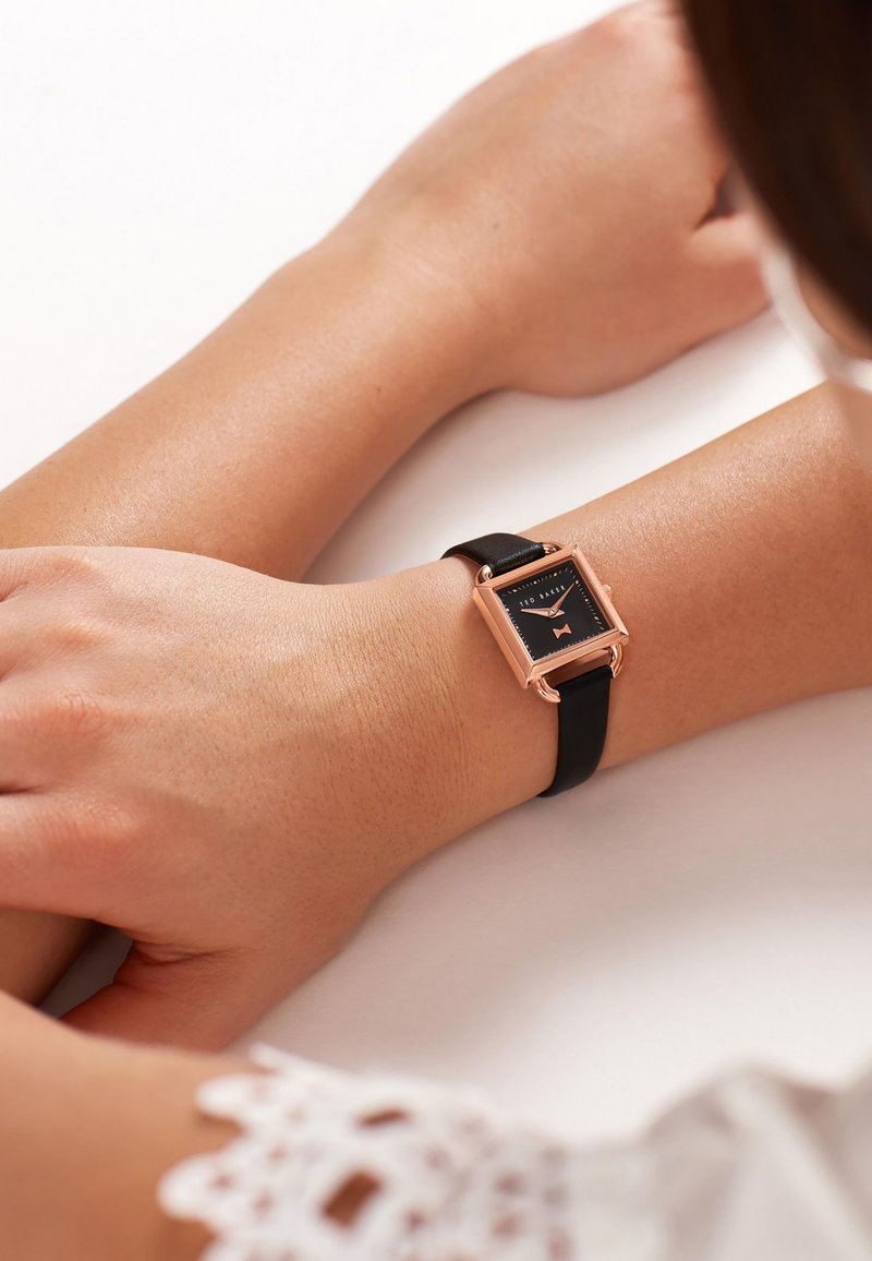 Montre rectangulaire avec un cadran noir, un cadre en or rose et un bracelet en cuir noir. Texture lisse avec des détails subtils sur le cadran.