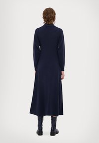Marineblaues Langarmkleid mit hohem Hals, in Midi-Länge, glatter Textur und voluminösem Schnitt. Getragen mit glänzenden kniehohen Stiefeln.