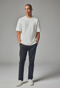 Weißes T-Shirt mit kurzen Ärmeln und Rundhalsausschnitt, kombiniert mit schwarzen Hosen und weißen Sneakers, vor einem schlichten grauen Hintergrund stehend.