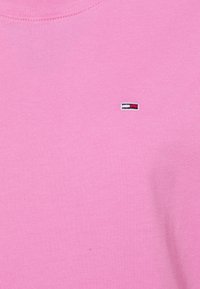 Tela rosa con un pequeño logotipo rectangular bordado en blanco, rojo y negro cerca de la parte central superior.