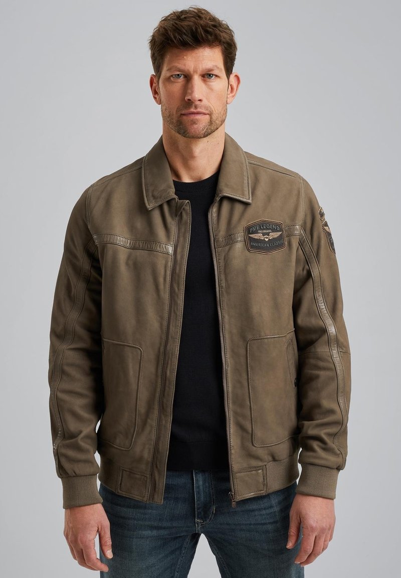 Veste bomber en cuir vert olive avec des poignets et un ourlet côtelés, des détails de patch sur la poitrine et des poches latérales, assortie à un jean foncé.