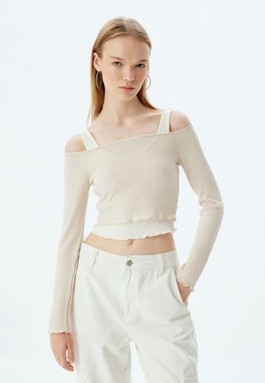 Lichtbeige geribde lange mouw top met een off-the-shoulder ontwerp, een cropped zoom en gerafelde randen. Model draagt witte high-waisted broek.
