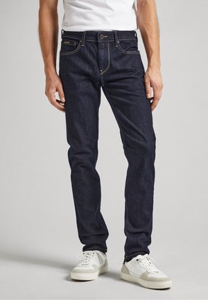 Mężczyzna ubrany w dopasowane ciemnoniebieskie dżinsy, białe sneakersy i białą koszulkę, stojący na tle jednolitego szarego tła.