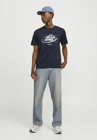 Marineblauwe katoenen t-shirt met wit grafisch tekstontwerp, gecombineerd met lichtblauwe denim jeans en grijze sneakers, accessoires met een lichtblauwe pet.