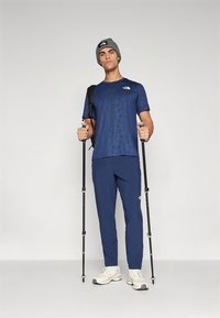 Camisola atlética azul-marinho com padrão geométrico, calças azul-marinho combinando, gorro cinzento e bastões de trekking. Ténis brancos completam o conjunto.