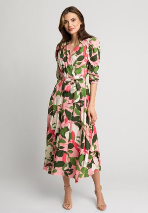 Kobieta stojąca na jednolitym tle w różowo-zielono-białej sukience kopertowej w kwiaty z bufiastymi rękawami i beżowych szpilkach na kostkę.