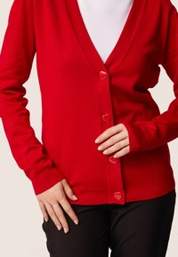 Cardigan rosso con scollatura a V, maniche lunghe e bottoni a forma di cuore. Il tessuto appare liscio con una silhouette aderente.