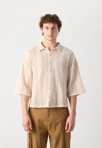 Beige kötött, rövid ujjú ing gombos elejével, texturált mintázattal és egyenes szegéllyel, khaki nadrággal párosítva.