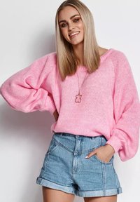 Różowy sweter z dzianiny o szerokich rękawach, zestawiony z podwiniętymi szortami dżinsowymi. Tekstura jest luźno dziergana, a kolory są żywe.