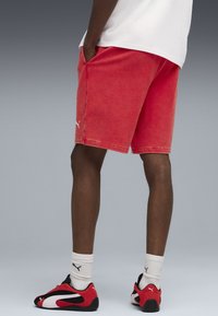 Mand iført røde shorts, hvid T-shirt, hvide Puma-sokker og sorte, røde og hvide Puma-sneakers, stående med hænderne i lommerne.