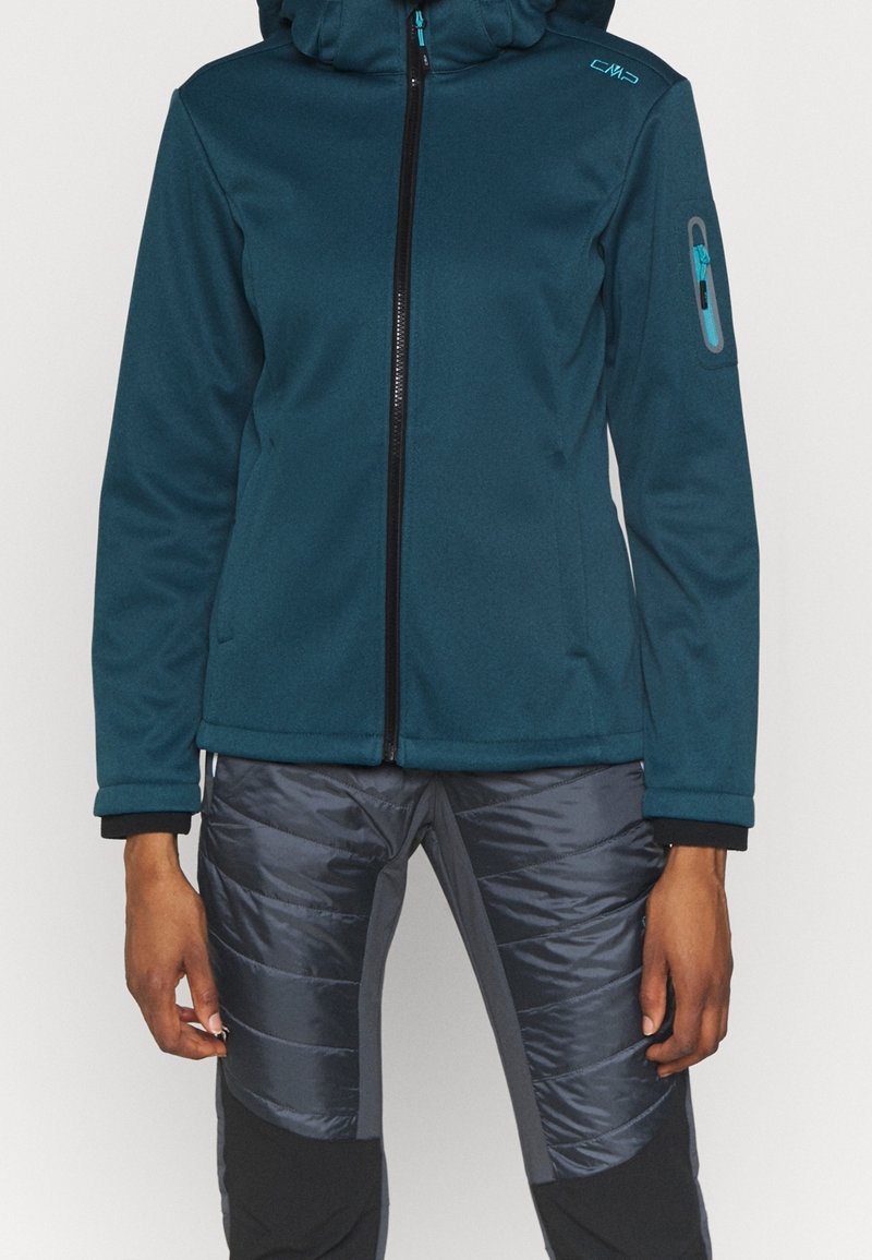 Veste bleu sarcelle à fermeture éclair avec capuche et poche sur le bras, associée à un pantalon matelassé isolé noir et gris sur une personne debout.