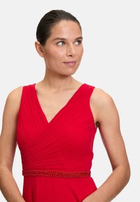 Rotes ärmelloses Kleid mit V-Ausschnitt, plissiertem Stoff und einem mit Perlen besetzten Taillenbund, der eine glatte Textur und eine lebhafte Farbe zeigt.