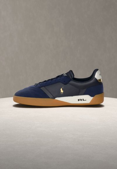 Zapatilla deportiva azul marino con suela de goma, lengüeta blanca en el talón, logo dorado de Polo en el lateral y letras "RL" en la media suela, mostrada sobre una superficie neutra.