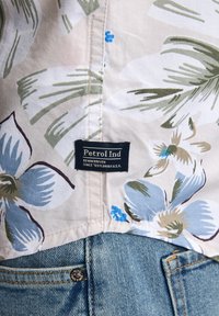 Camisa de manga corta con estampado floral sobre un fondo beige, con flores azules y hojas verdes, y una etiqueta negra "Petrol Ind" sobre unos pantalones cortos de mezclilla.