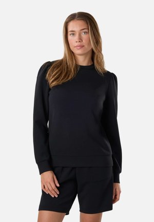 Vrouw met lang haar die een zwarte top met lange mouwen met opgepofte schouders en zwarte korte broek draagt, staand tegen een witte achtergrond.