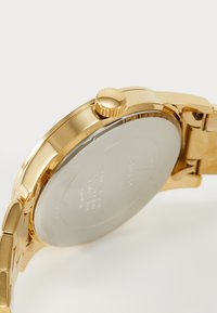 Orologio in metallo dorato con cassa rotonda, corona texturizzata e bracciale a maglie. Fondello in argento con dettagli incisi. Finitura liscia, superficie riflettente.