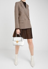 Valkoinen rakenteellinen käsilaukku, jossa on pyöreä kultasulkija, mustat yksityiskohdat, yhdistettynä ruskeaan kimaltelevaan mekkoon ja haukansilmätakkiin.