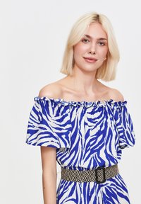 Kobieta z blond włosami do ramion nosi niebiesko-białą sukienkę w paski zebry, odkrytą na ramionach, z przewiązanym czarnym paskiem w talii.