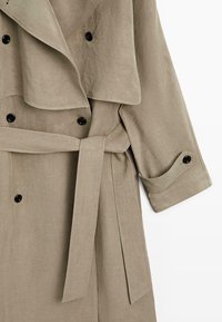 Beige trenchcoat med sorte knapper, bred krave, bælte bundet i taljen og knaplukket manchet på lang ærme, vist på hvid baggrund.