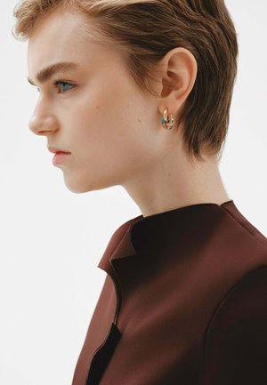 Pendientes de aro de oro con un acento multicolor, con un diseño circular. La modelo lleva una blusa marrón con un cuello estructurado.