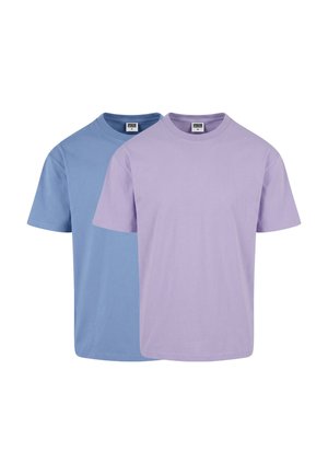 Jeune homme aux cheveux courts portant un T-shirt uni bleu clair à manches courtes et un pantalon foncé, debout devant un fond gris.