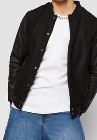 Svart bomberjacka med ull-kropp och lädersleeves, med tryckknappar, rund krage, bärs över en vit t-shirt och blå denim.