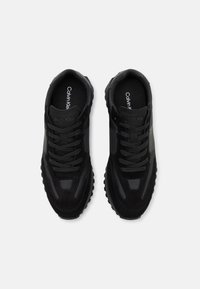 Black athletic shoes with a mixed material design featuring suede and mesh. The laces are simple, and the sole is textured for grip.