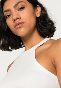 Top branco canelado sem mangas com gola alta, apresentando um design ajustado e uma textura suave, destacando uma estética limpa e minimalista.