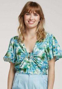 Blouse florale dans des tons de bleu, vert et rose. Présente un décolleté en V profond, des manches évasées et un détail froncé à l'avant avec un petit accent en métal.