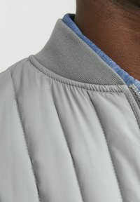 Giacca trapuntata grigia con colletto grigio a coste e fodera in tessuto blu, caratterizzata da cuciture diagonali e una texture liscia e lucida.