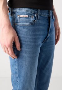 Tuvredzēts tuvplāks, kurā redzams cilvēks, kurš valkā zilos Calvin Klein džinsus ar redzamu zīmi uz priekšējā kabatas, rokas dabiski noliktas blakus.