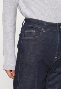 Dark blue denim jeans with gold stitching, featuring a side pocket and a high waistband. Paired with a light gray knitted sweater.