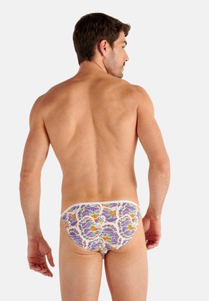 Male model facing away wearing white briefs with a colorful purple, orange, and green floral pattern on a plain background.