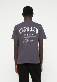 T-shirt de algodão cinza com grande texto gótico branco nas costas, com gráficos adicionais e um logotipo abaixo. Mangas curtas e decote redondo.