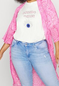 En person bär en vit t-shirt med en ögongrafik och texten "COSMIC VISION", tillsammans med en rosa mönstrad kimono och blå jeans.