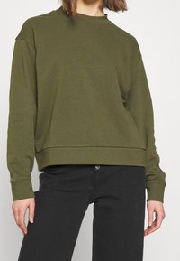 Sudadera verde oliva con cuello redondo, hombros caídos y puños acanalados. Combinada con jeans negros de textura suave.